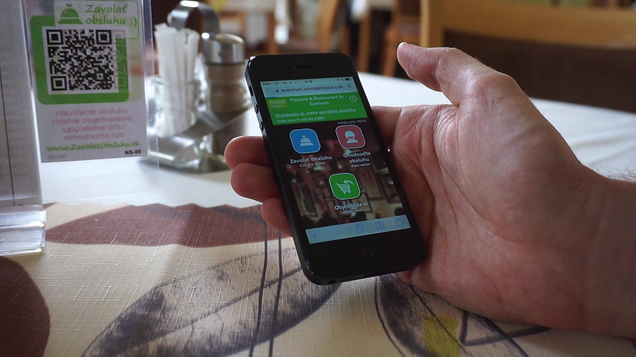 Hosť drž&iacute; smartf&oacute;n s otvorenou aplik&aacute;ciou na objedn&aacute;vanie pri stole &mdash; na obrazovke s&uacute; tlačidl&aacute; Zavolať obsluhu, Ohodnotiť obsluhu a Objednajte si, v pozad&iacute; QR k&oacute;d stojan
