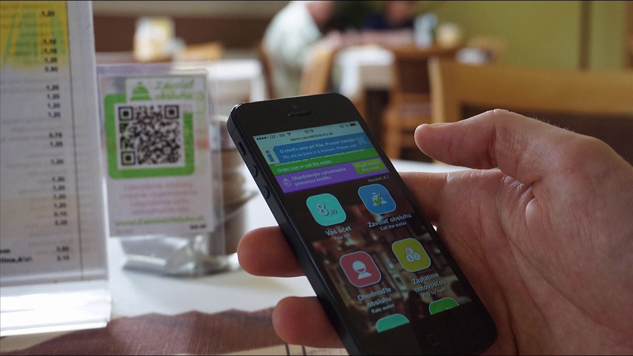 Smartf&oacute;n na re&scaron;tauračnom stole zobrazuje aplik&aacute;ciu iKelp so z&aacute;kazn&iacute;ckou obrazovkou &mdash; V&aacute;&scaron; &uacute;čet, Zavolať obsluhu, Ohodnotiť obsluhu, Zaplaťte hotovosťou a možnosť objedn&aacute;vky